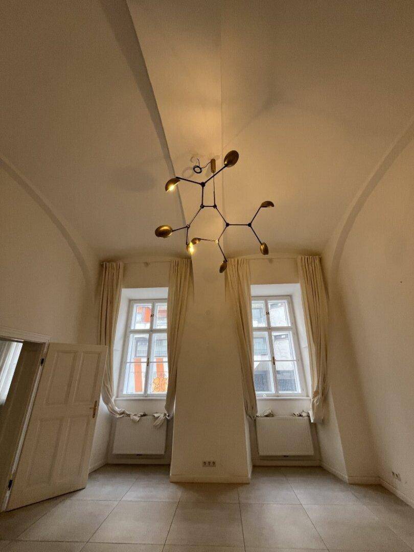 Geräumiger Wohnraum mit hohen Gewölbedecken, einem modernen Kronleuchter und zwei großen Fenstern.