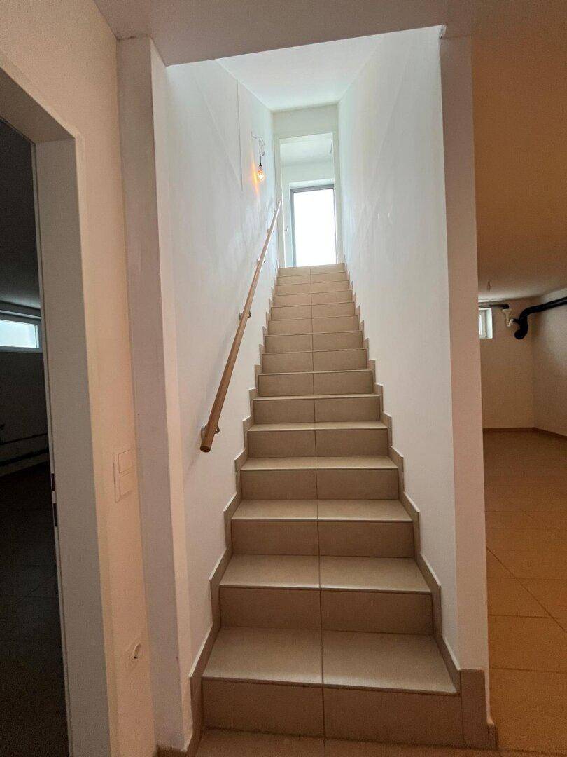 Heller Treppenaufgang mit Fliesenstufen und Holzhandlauf, der nach oben führt.