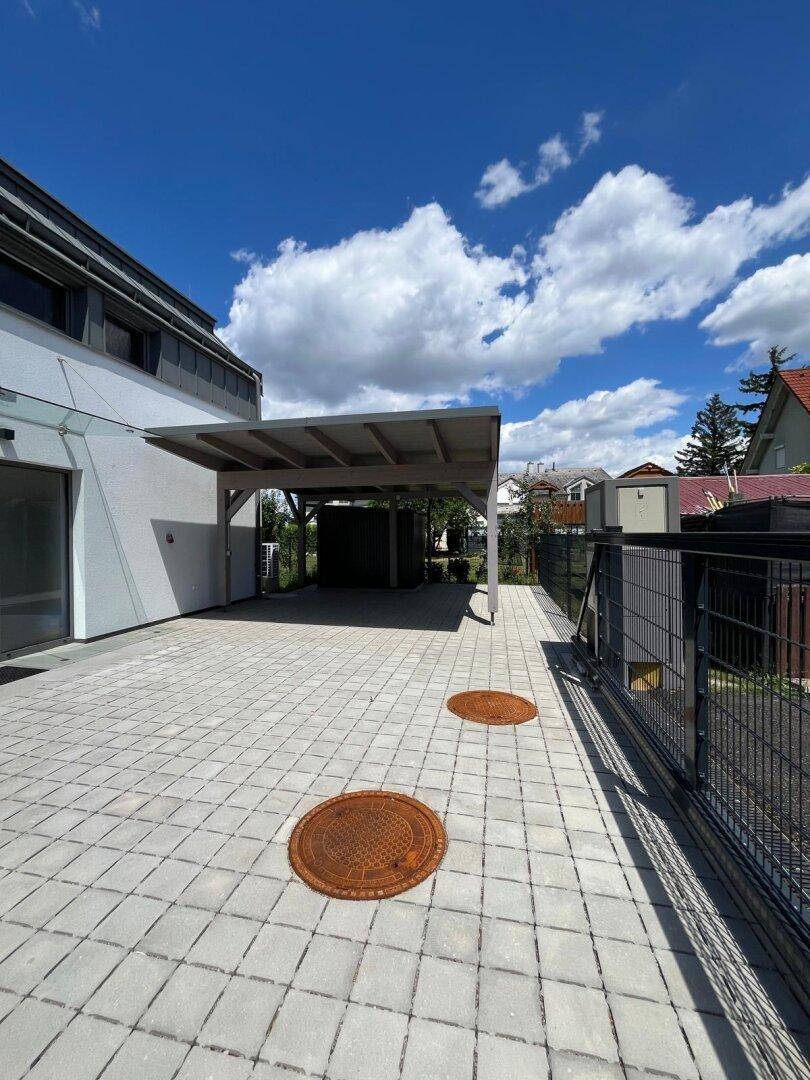 Seitenansicht des Hauses mit Carport, gepflastertem Bereich und Zaun unter blauem Himmel.
