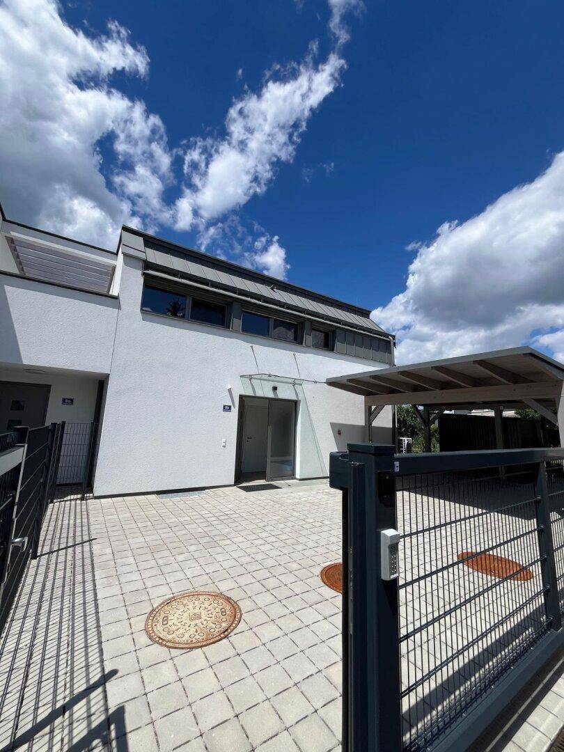 Modernes Reihenhaus mit weißer Fassade, Carport und gepflastertem Eingangsbereich unter blauem Himmel.