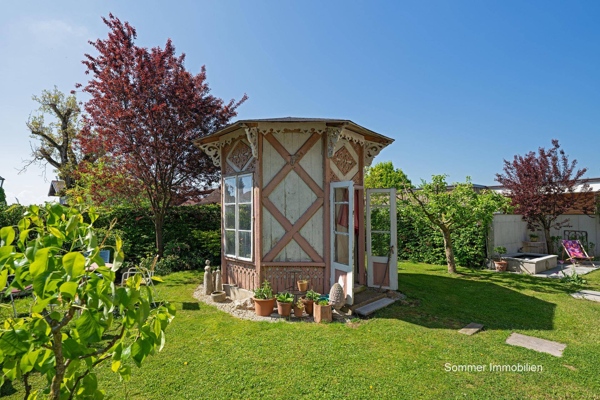 Malerischer Garten mit einem dekorativen Gartenhaus, umgeben von Bäumen und grüner Vegetation.