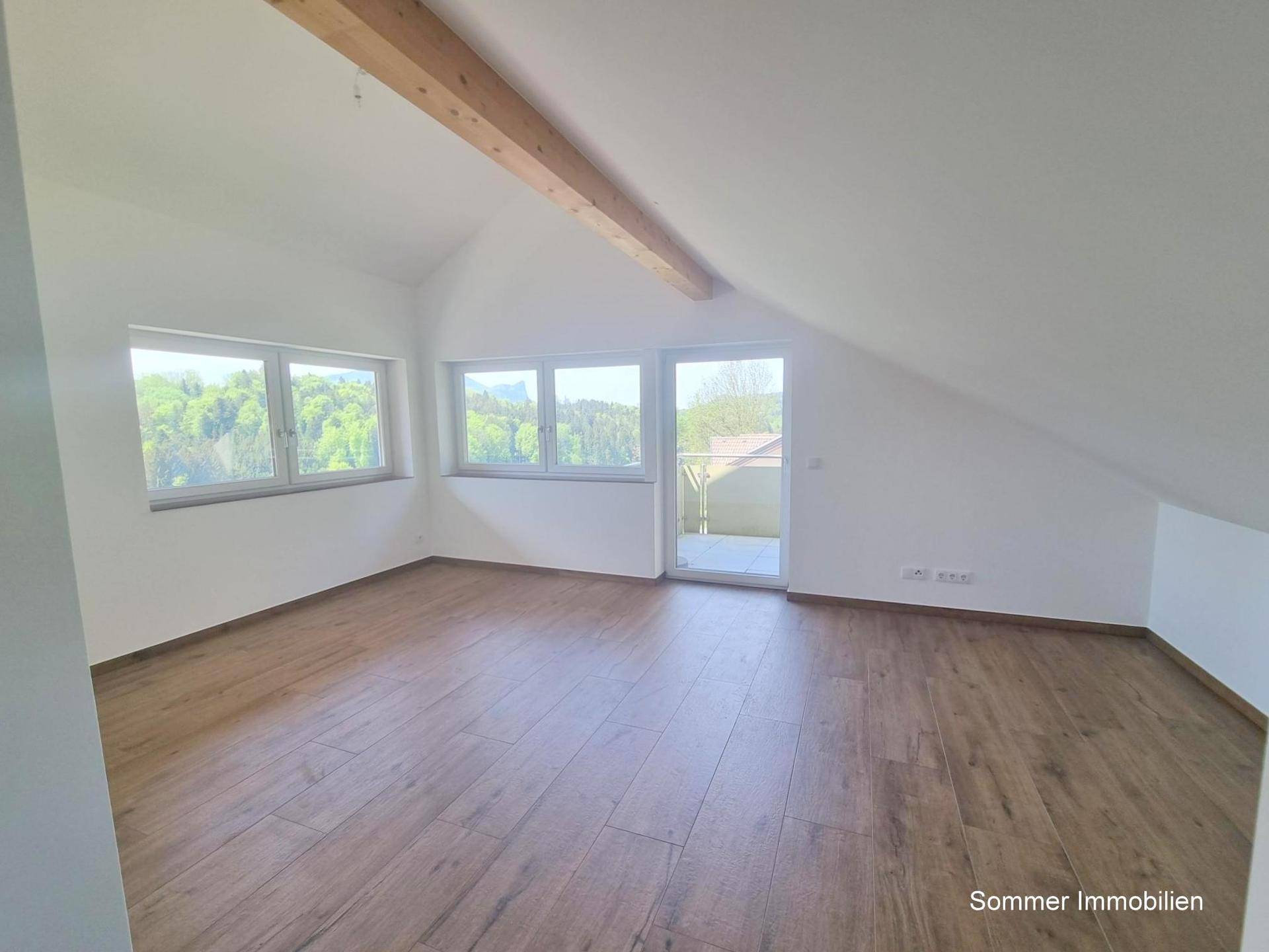 Geräumiger Raum mit Holzboden, großen Fenstern und Zugang zu einem Balkon, unter einer Dachschräge.