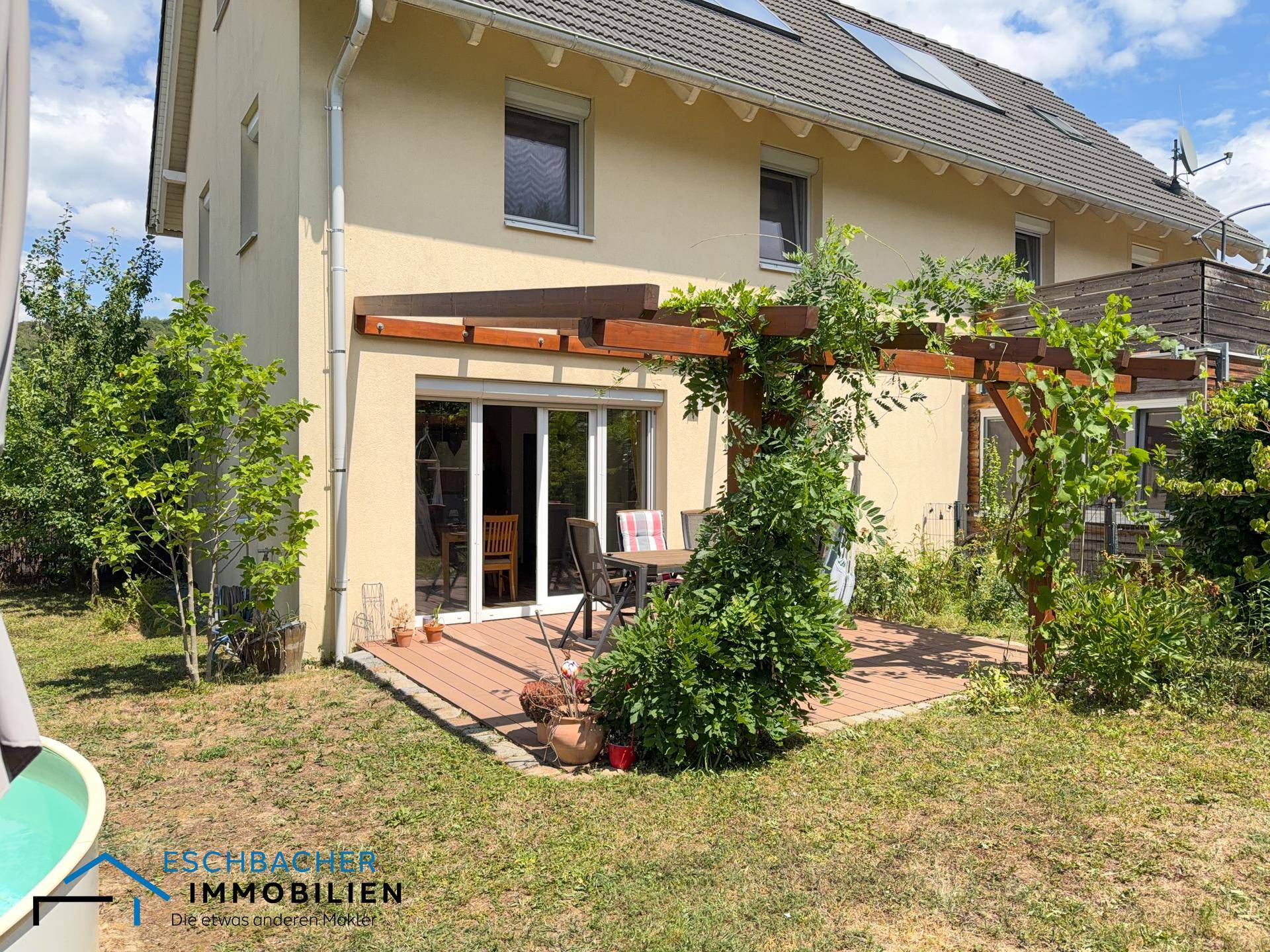 Terrasse am Haus mit Holzpergola und Gartenmöbeln, umgeben von Grünflächen.