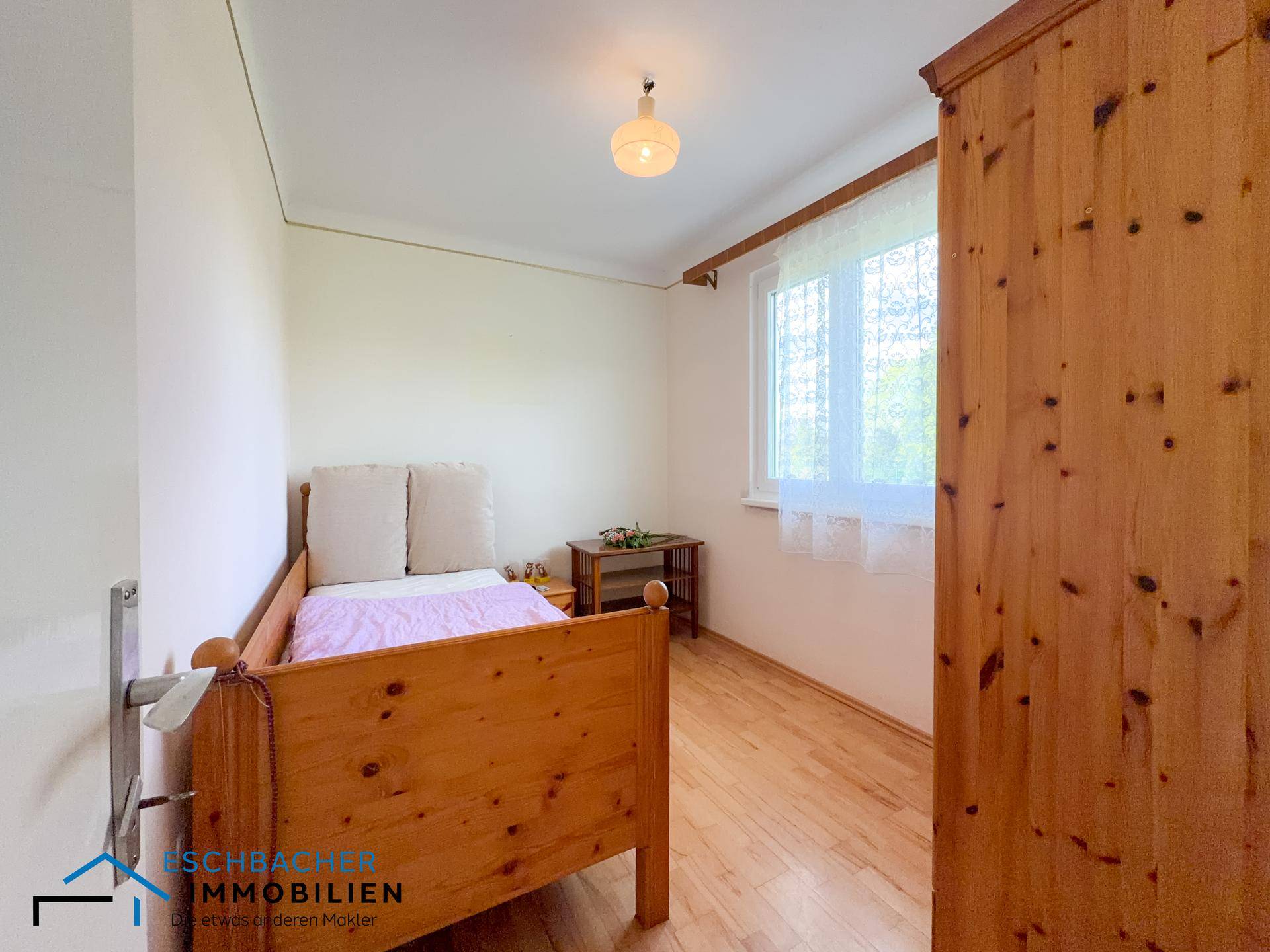 Helles Schlafzimmer mit Holzbett, Kleiderschrank und Fenster mit Vorhängen.