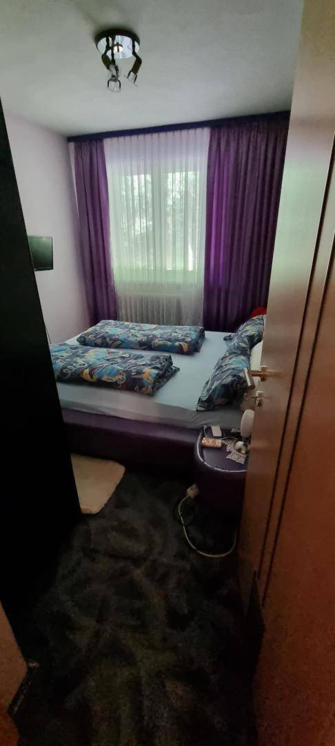 Schlafzimmer mit Doppelbett, lila Vorhängen und Teppichboden, seitlich aufgenommen.