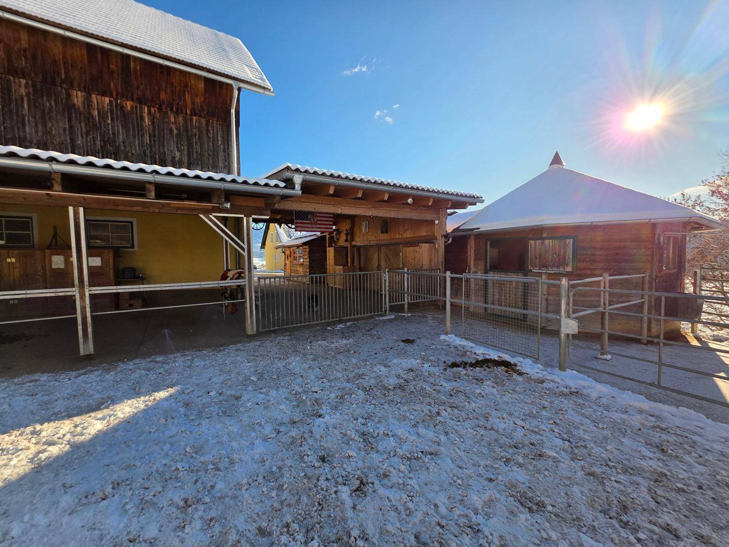 Außenbereich eines Stalls mit Holzzäunen und einem überdachten Unterstand, teilweise mit Schnee bedeckt.