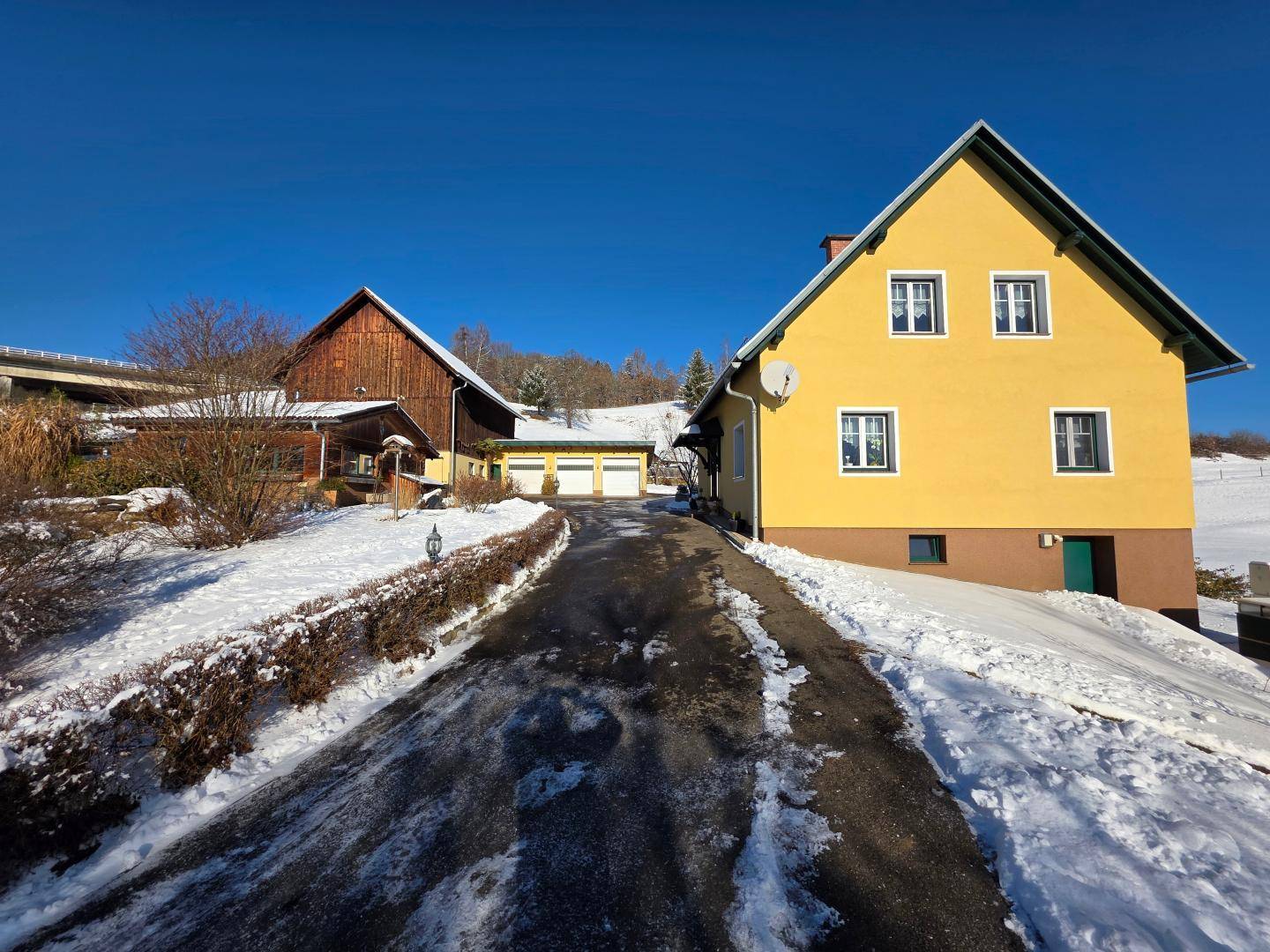 Gelbes Wohnhaus mit braunem Sockel und einem Nebengebäude, umgeben von Schnee an einem sonnigen Wintertag.