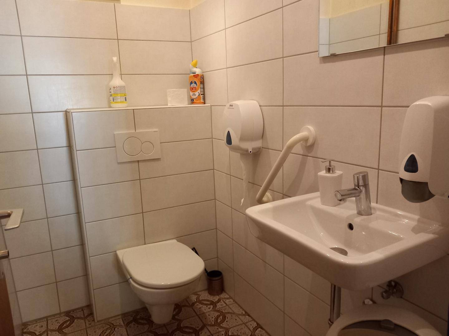 Gefliestes Badezimmer mit wandhängender Toilette, Waschbecken und Spendern für Seife und Papier.