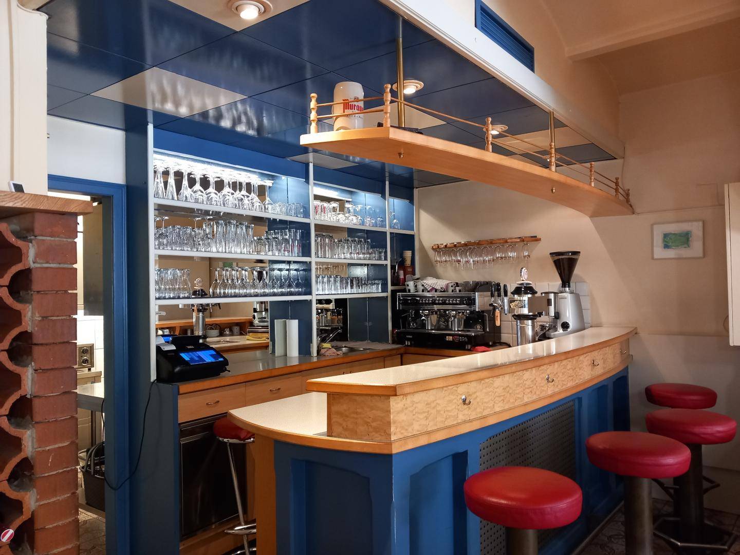 Detailansicht der Bar mit Kaffeemaschine, Gläserregalen und roten Barhockern, bereit für den Service.