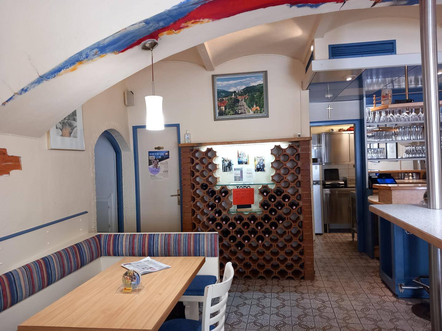 Sitzbereich im Café mit gepolsterten Bänken, einem dekorativen Weinflaschenregal und Zugang zu weiteren Räumen.