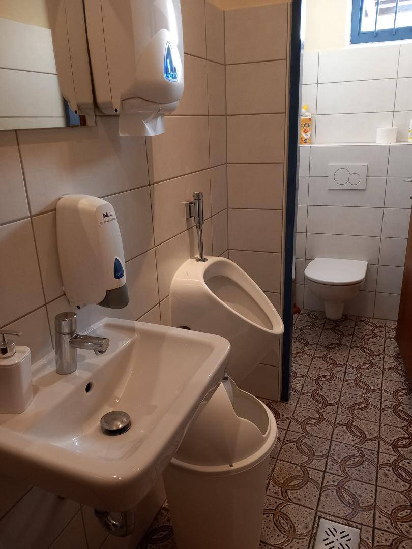 Toilettenraum mit Waschbecken, Urinal und WC, alles in weißen Fliesen gehalten und mit einem Fenster versehen.