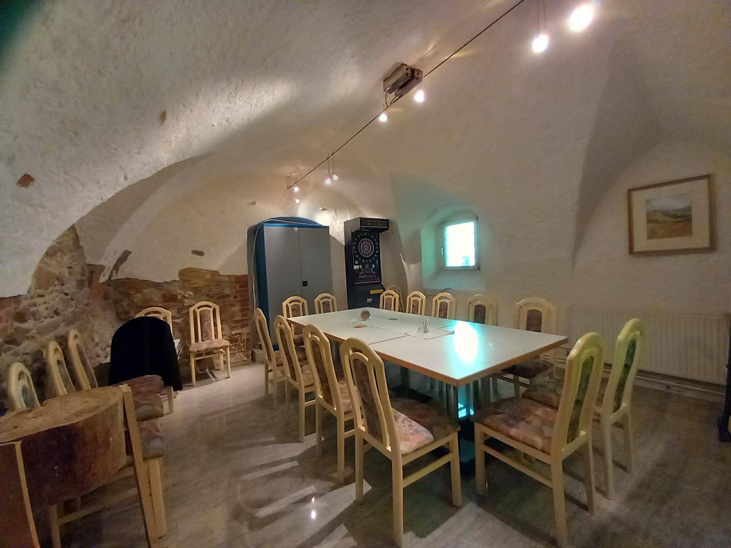 Gewölbekeller mit einem großen Tisch, Stühlen und einer Dartscheibe, ideal für Versammlungen.