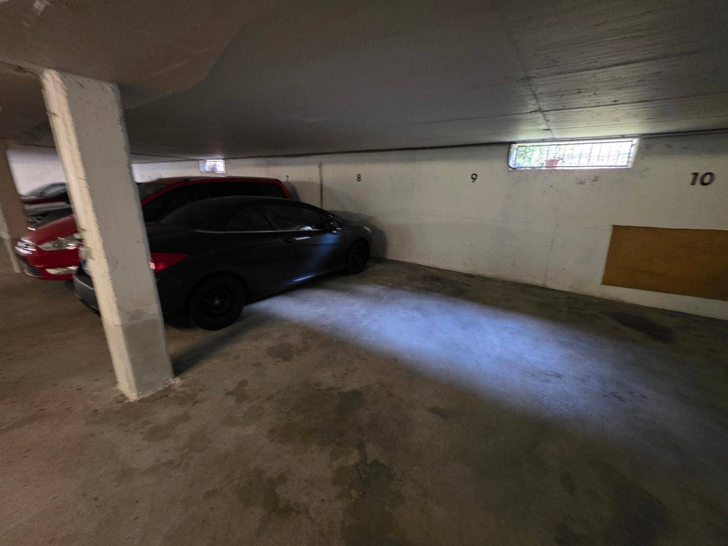 Tiefgarage mit geparkten Autos und Betonstützen, beleuchtet durch ein kleines Fenster.