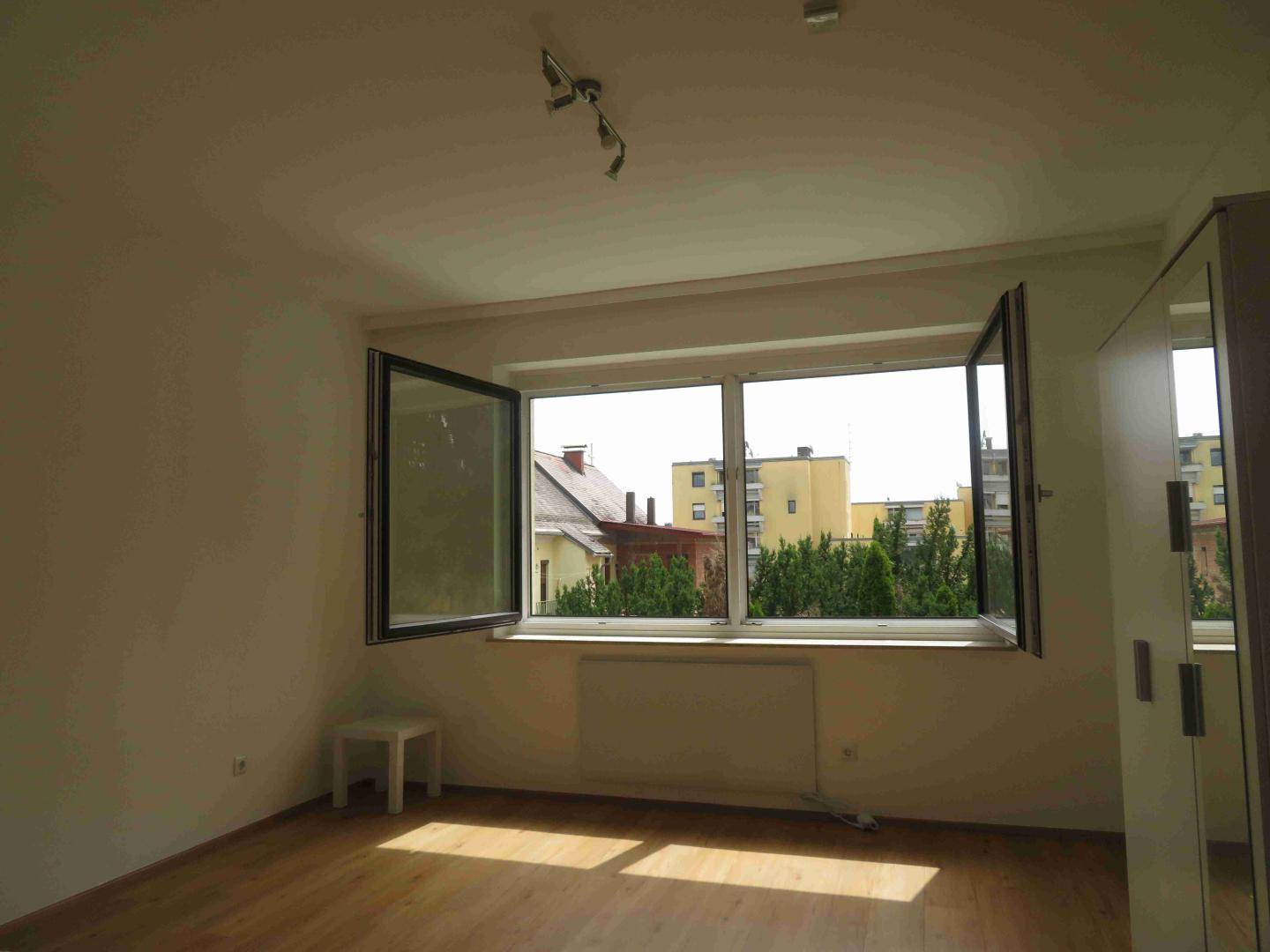Helles Schlafzimmer mit Holzboden und großen Fenstern, die viel Tageslicht hereinlassen.