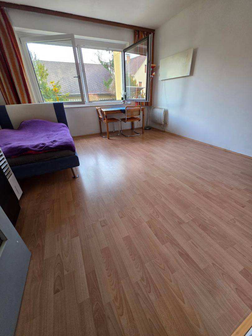 Wohnbereich mit Holzboden, Bett und kleinem Esstisch, mit geöffnetem Fenster für frische Luft.
