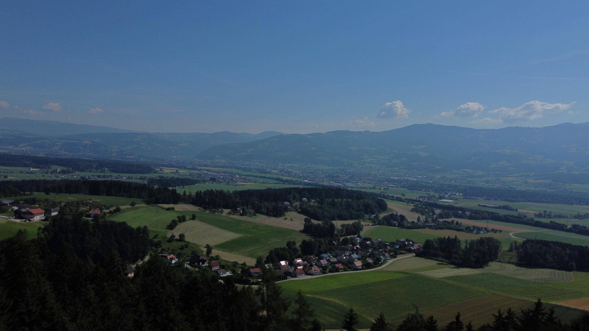 Weitläufige Luftaufnahme eines Tals mit Feldern, Wäldern und einer kleinen Siedlung unter blauem Himmel.