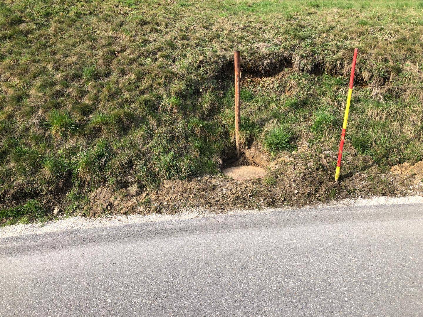 Nahaufnahme eines grasbewachsenen Hangs neben einer Straße mit einem Kanaldeckel und Markierungspfählen.