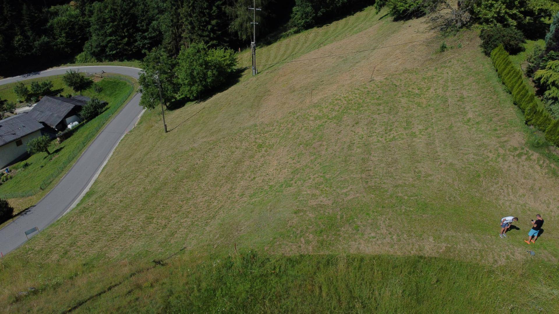 Luftaufnahme eines großen, grünen Grundstücks neben einer kurvigen Straße mit zwei Personen am Rand.