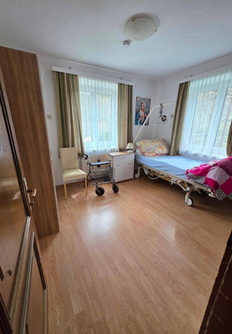 Schlafzimmer mit einem Pflegebett, Holzschrank und zwei Fenstern, die für viel Tageslicht sorgen.