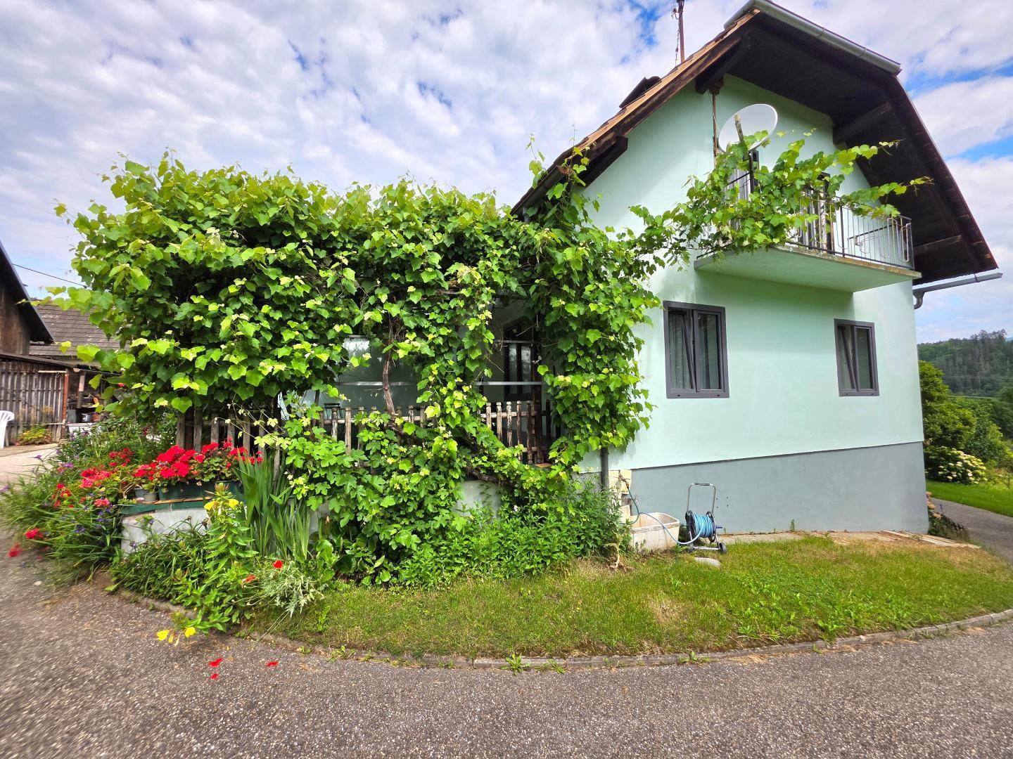 Hausfassade, teilweise von Weinreben bewachsen, mit bunten Blumenbeeten und einer Zufahrt.