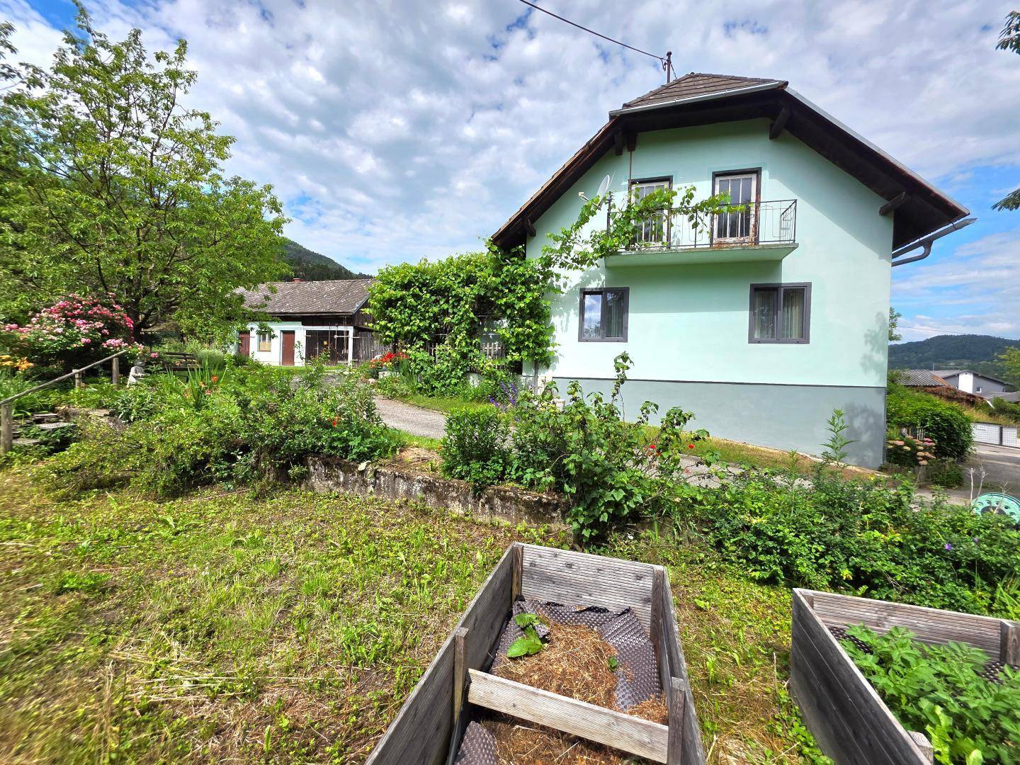 Gartenbereich mit Hochbeeten und Rasenfläche vor dem hellgrünen Haus, umgeben von üppiger Natur.