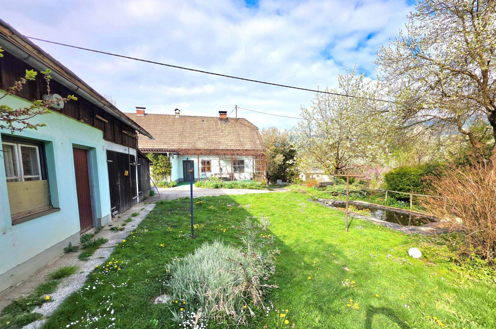 Gartenlandschaft mit einem kleinen Teich, weitläufigem Rasen und Nebengebäuden im Hintergrund unter blauem Himmel.