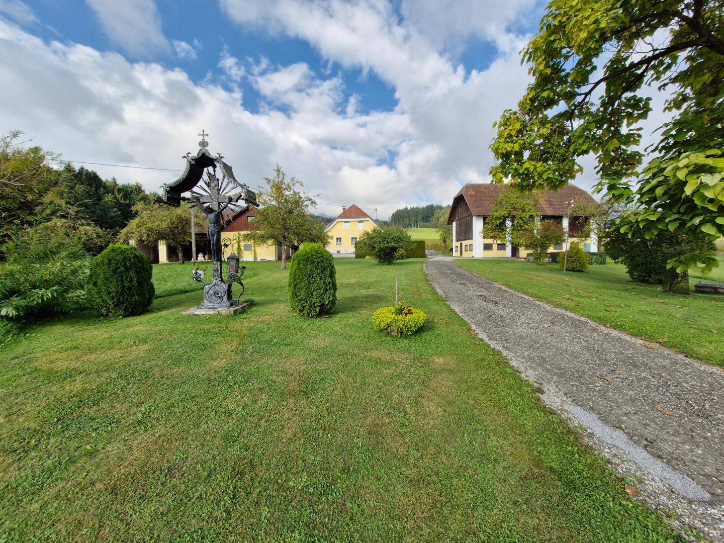 Weitläufiges Anwesen mit mehreren Gebäuden, langer Auffahrt, grünem Rasen und einem religiösen Denkmal in ländlicher Umgebung.