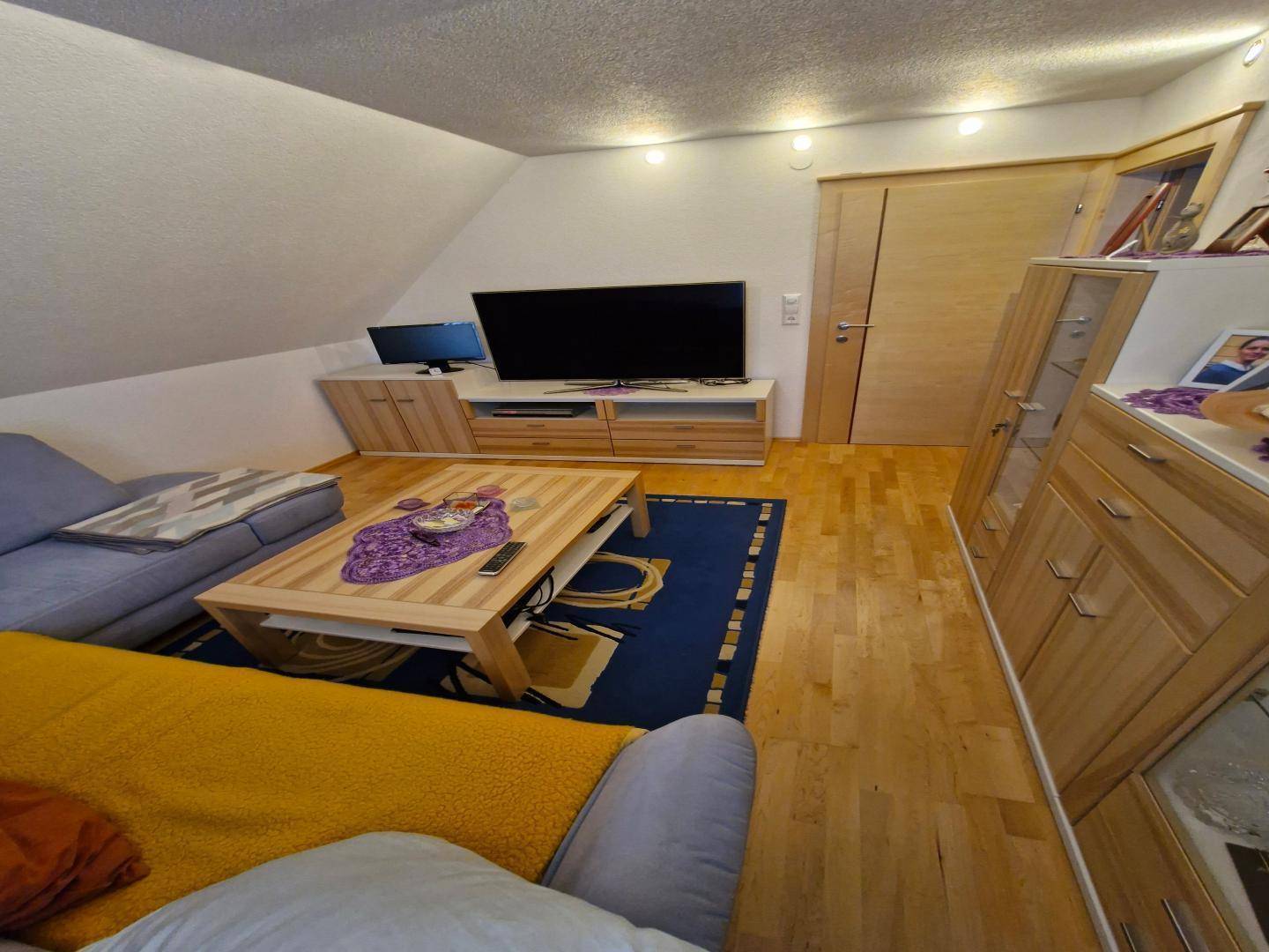 Wohnzimmer im Dachgeschoss mit Sofa, Couchtisch, Fernseher und Holzschränken unter der Dachschräge.