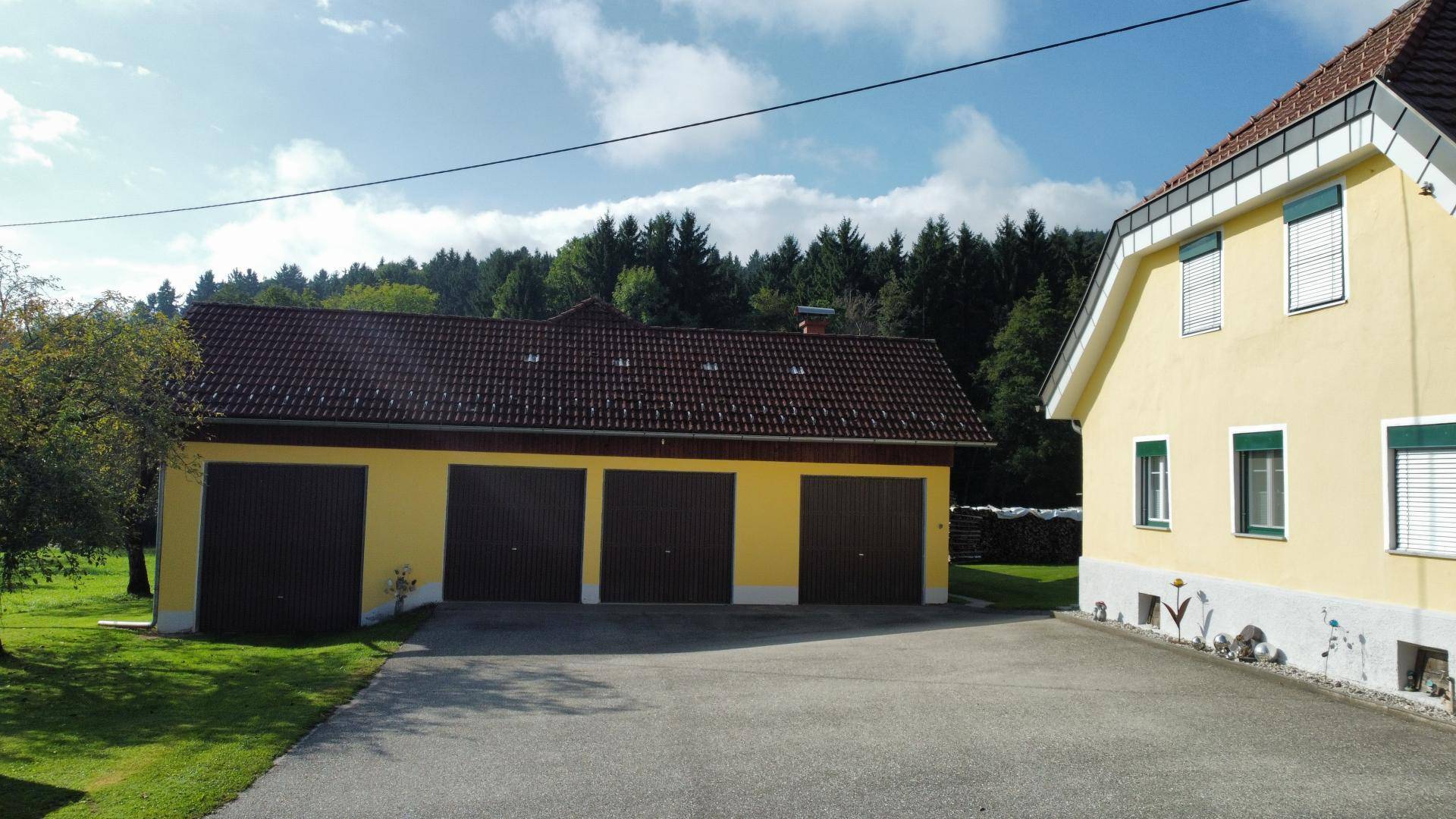 Gelbes Wohnhaus und separates Garagengebäude mit drei Toren, umgeben von Bäumen und einer asphaltierten Zufahrt.