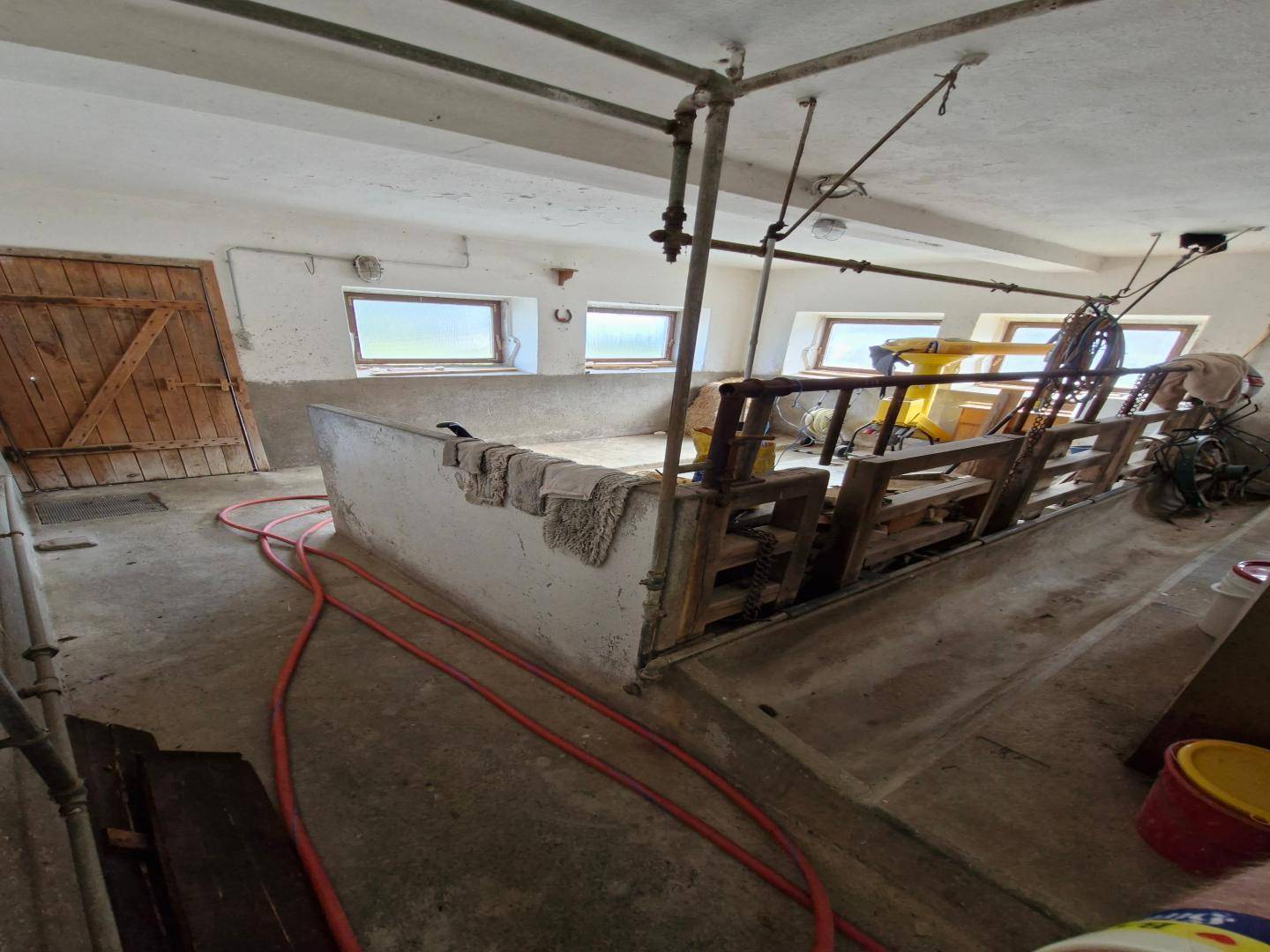 Innenansicht einer Scheune oder eines Stalls mit Betonboden, kleinen Fenstern und landwirtschaftlichen Geräten.