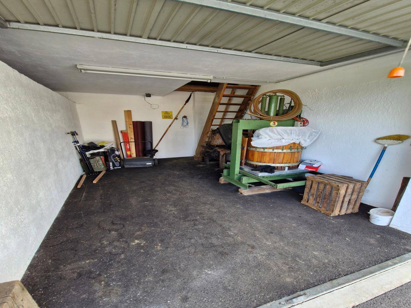Großer Garagenraum mit Betonboden, einer Holzpresse und einer Holztreppe, bietet viel Stauraum.