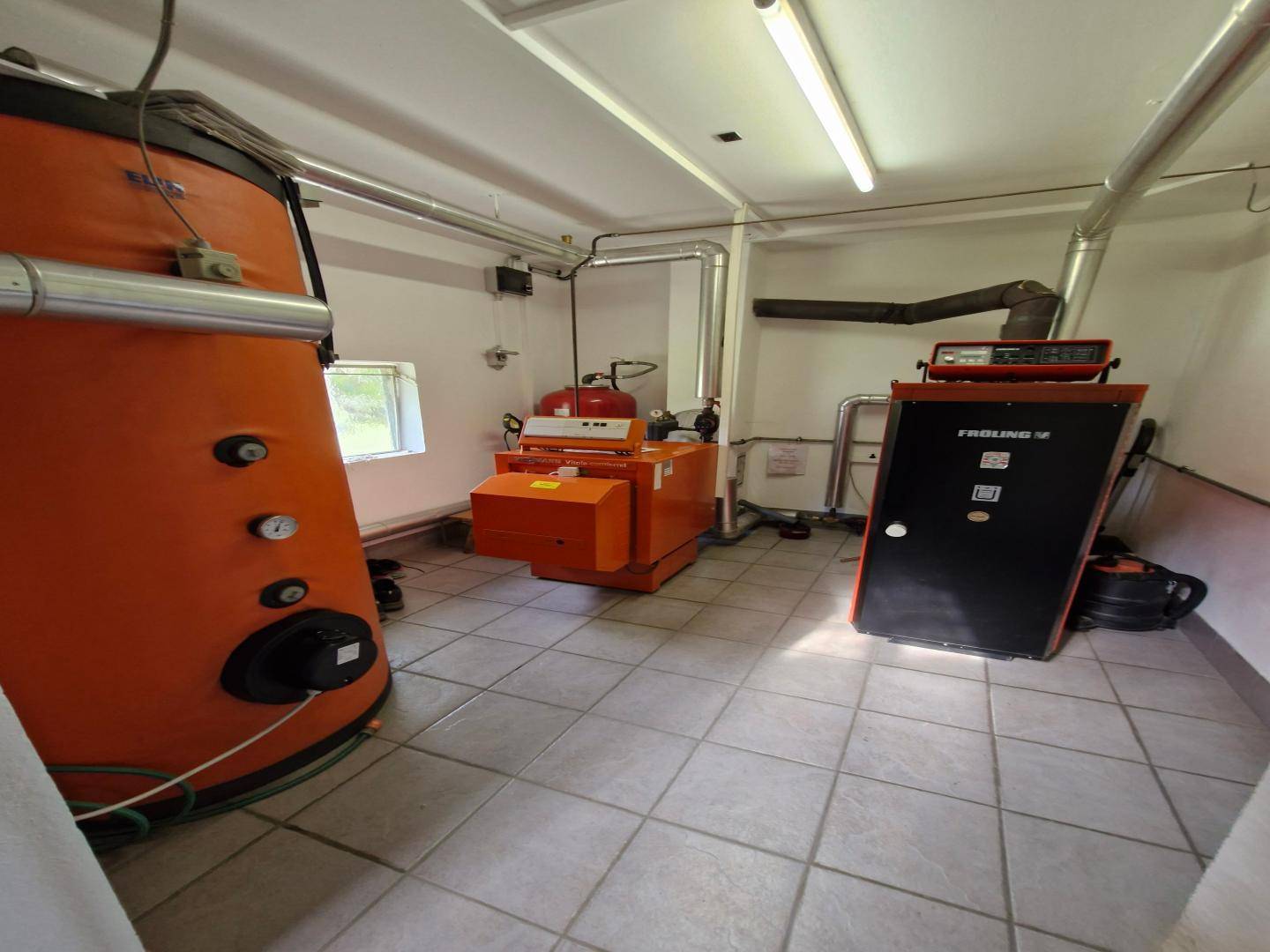 Heizungsraum mit großen orangefarbenen Heizkesseln, Rohren und Fliesenboden, gut gewartet und funktionsfähig.