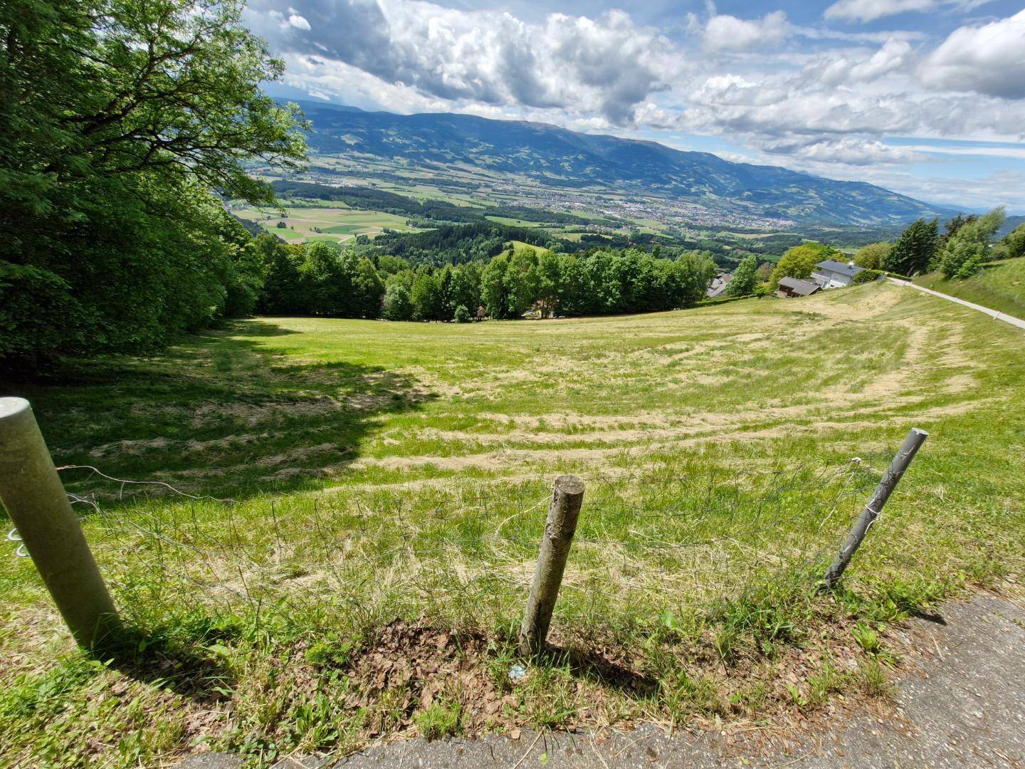 Blick über eine grüne Wiese mit sanften Hügeln und einem weiten Tal im Hintergrund unter bewölktem Himmel.