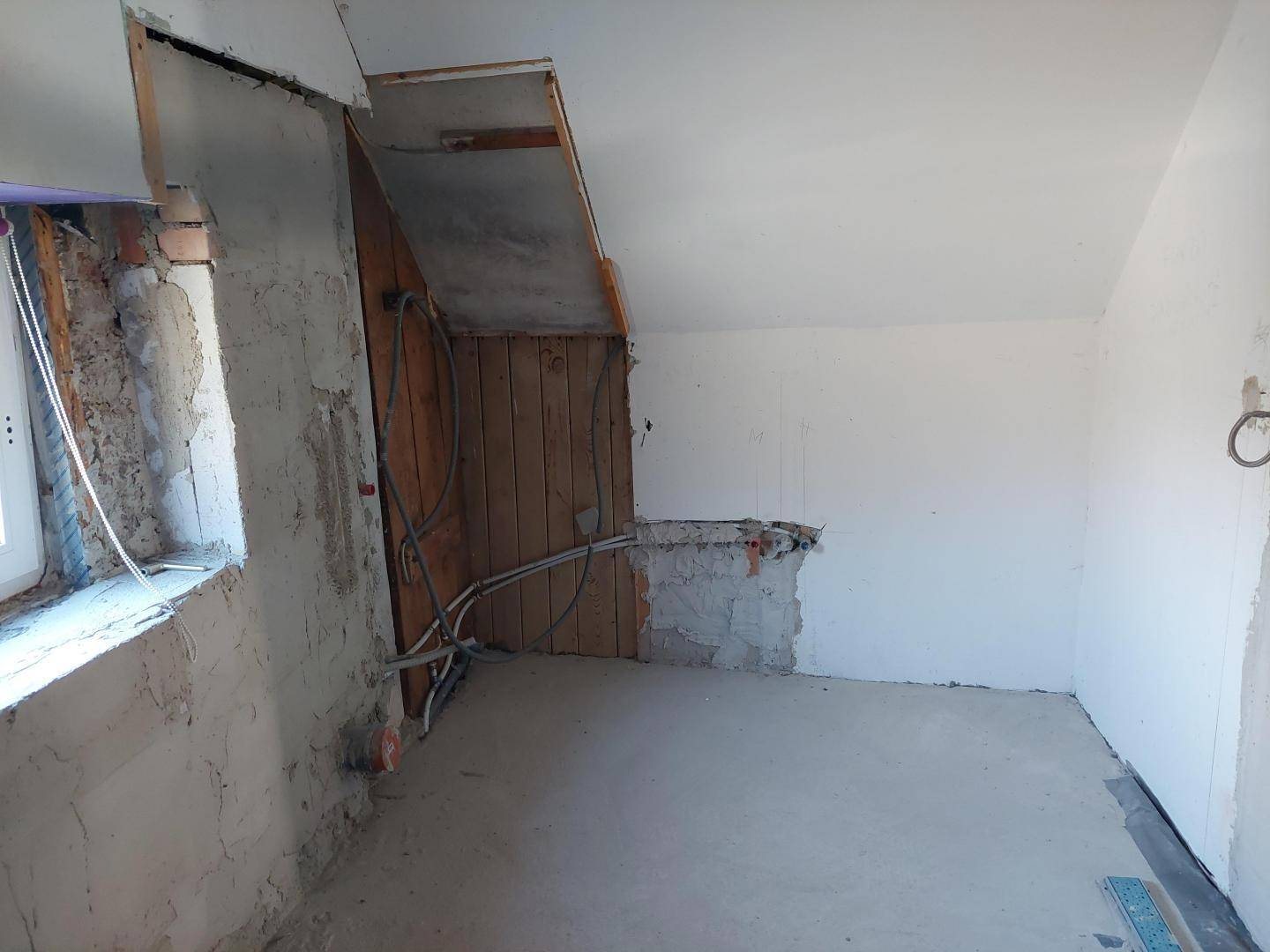Dachgeschossraum im Rohbau mit freiliegenden Leitungen und schrägen Wänden.