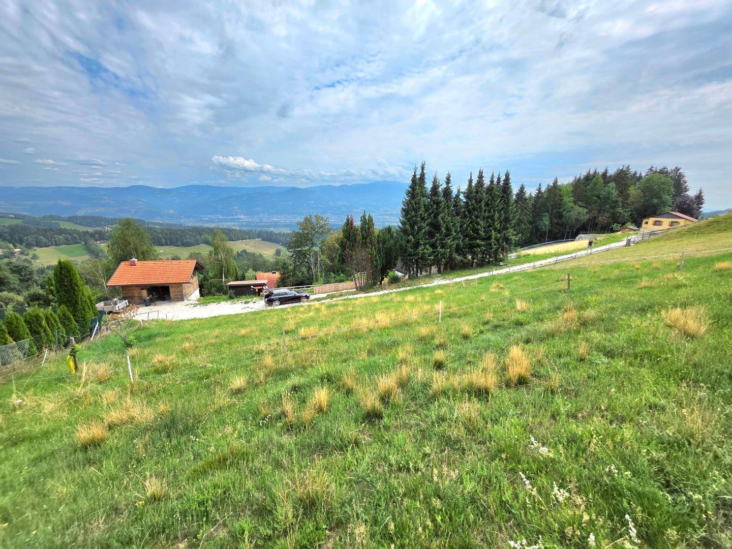 Panoramablick über ein grünes Grundstück mit Häusern und Bergen in der Ferne unter bewölktem Himmel.