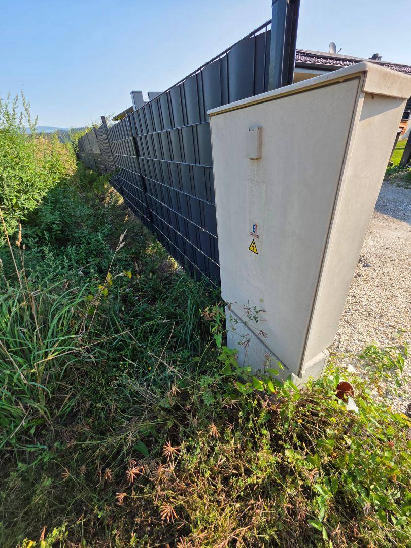 Grauer Stromkasten neben einem Zaun und einem Schotterweg in ländlicher Umgebung.
