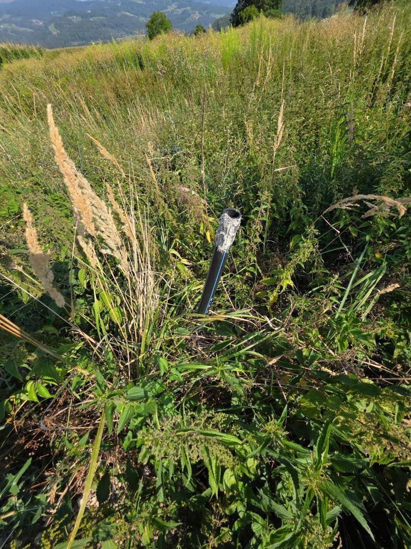Schwarzes Rohr ragt aus hohem Gras hervor, mit hügeliger Landschaft im Hintergrund.