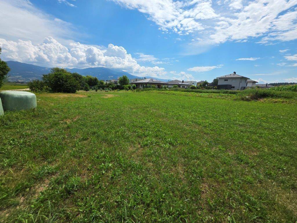 Großes, offenes Feld mit frischem Gras, umgeben von Häusern und Bergen am Horizont.