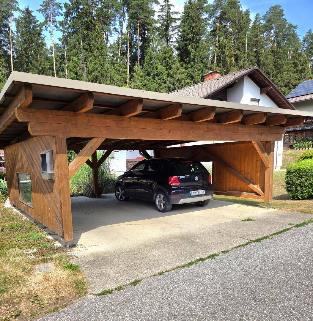 Holzcarport mit einem geparkten Auto und Wald im Hintergrund an einem sonnigen Tag.