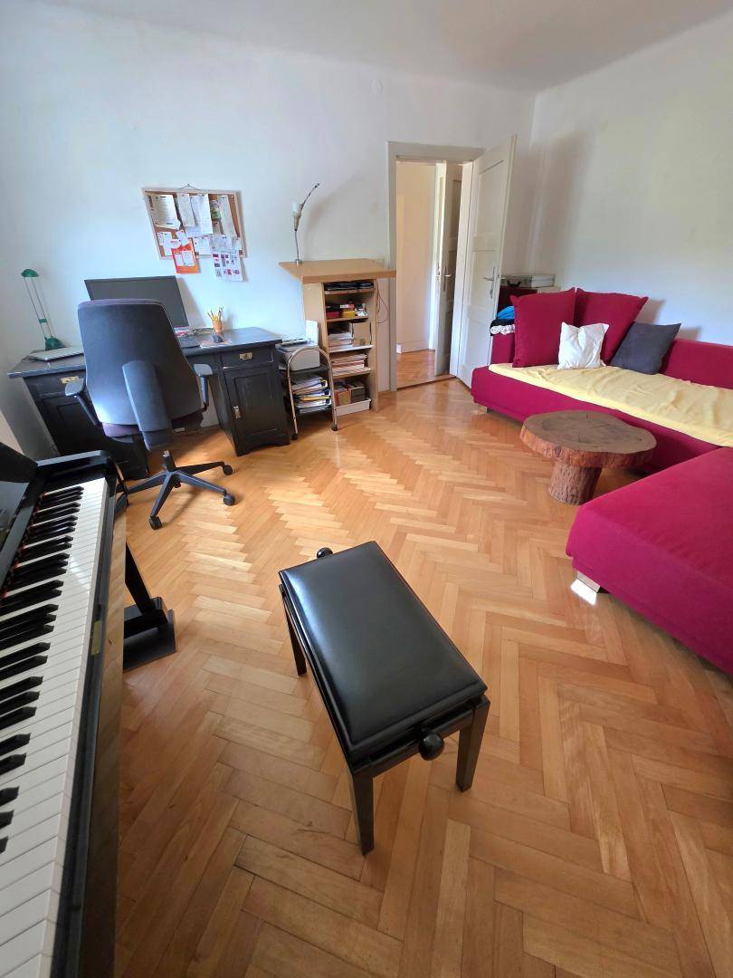 Geräumiges Wohnzimmer mit Fischgrätparkett, Klavier, Schreibtisch und rotem Sofa, hell und gemütlich.