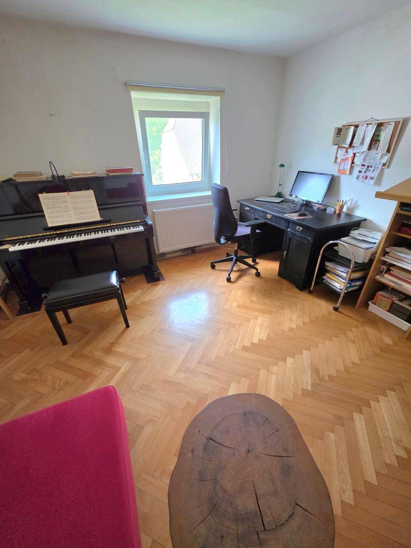 Helles Wohnzimmer mit Klavier, Schreibtisch und großem Fenster, ausgestattet mit Fischgrätparkett.