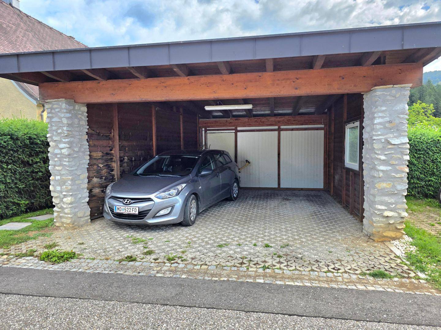 Überdachter Carport mit Holzbalken und Steinsäulen, bietet Schutz für ein Fahrzeug.
