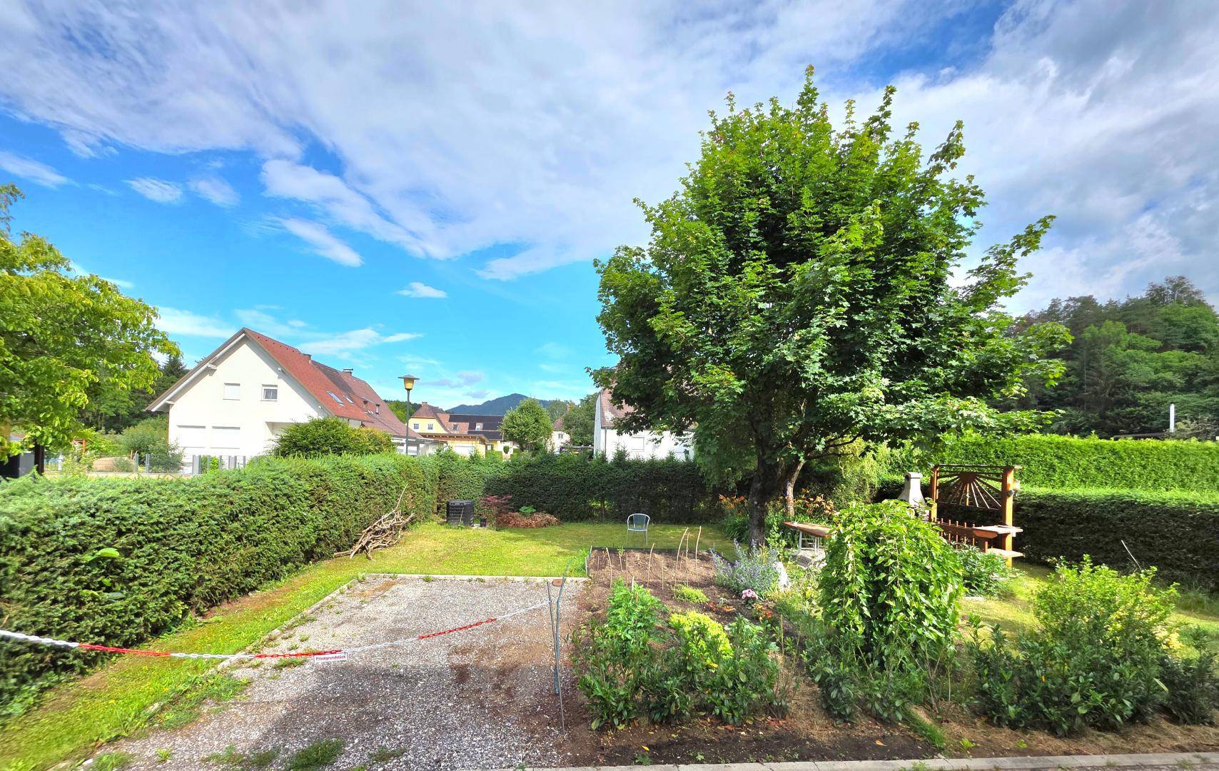 Grüner Gartenbereich mit hohen Hecken, Bäumen und Blick auf benachbarte Häuser unter blauem Himmel.
