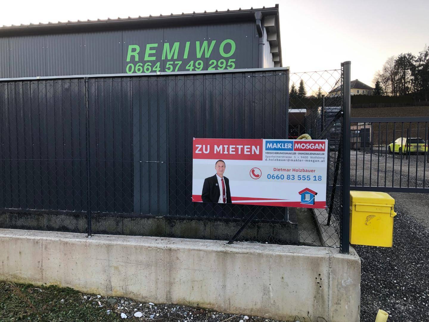 Gebäudeaußenansicht mit Firmenlogo und einem 'Zu vermieten'-Schild an einem Metallzaun mit gelbem Briefkasten.