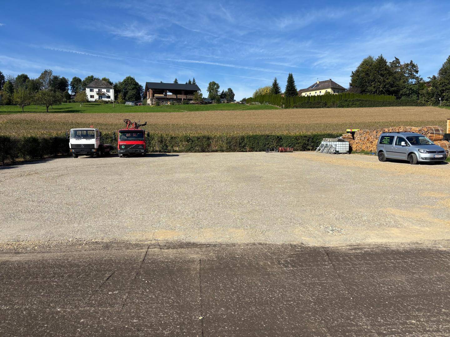 Weitläufiger Schotterparkplatz mit mehreren Fahrzeugen und Holzstapeln vor einer ländlichen Landschaftskulisse.