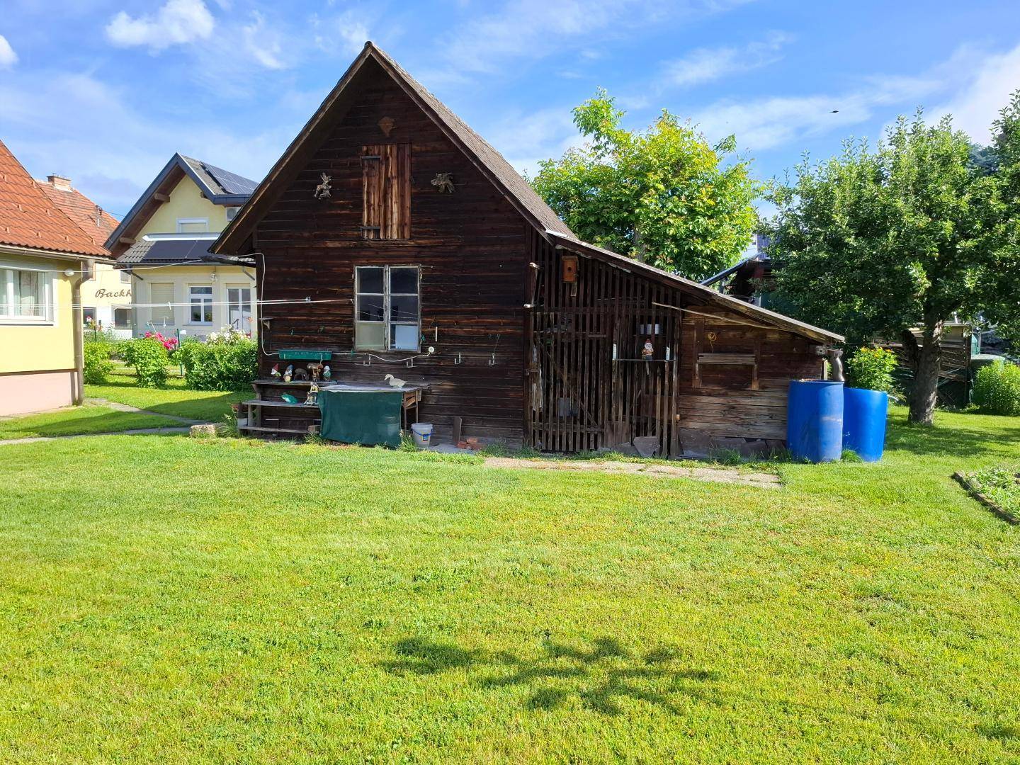 Rustikales Holzgebäude im Garten mit Rasenfläche und Bäumen unter blauem Himmel.