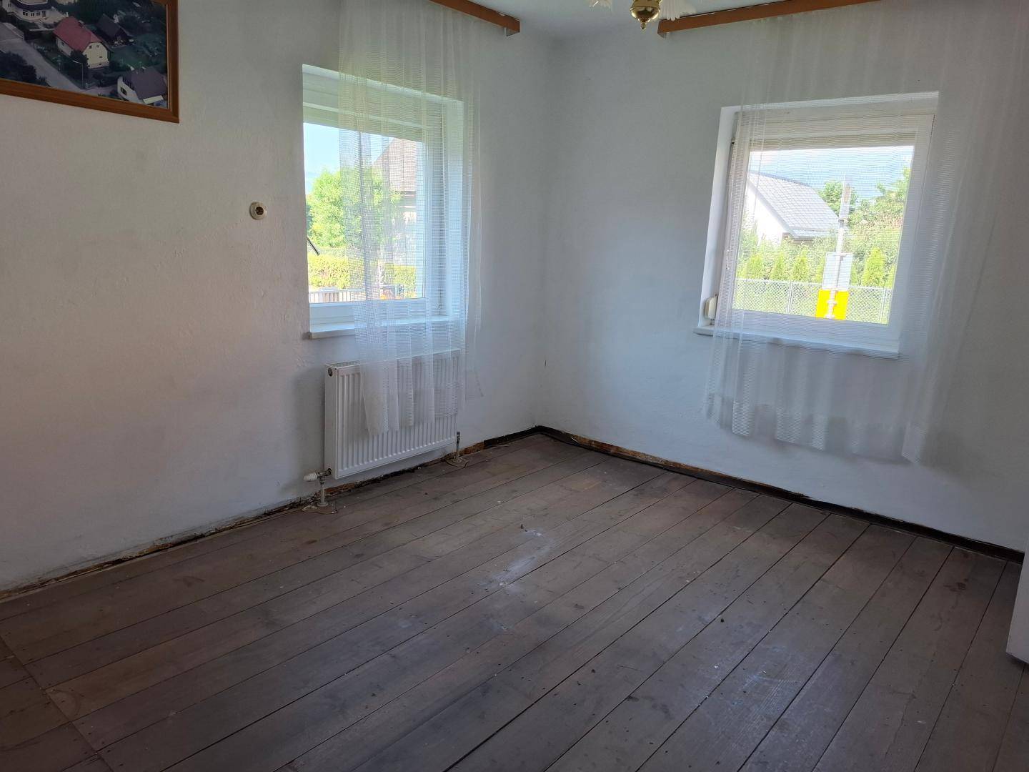 Leeres Zimmer mit Holzdielenboden, zwei Fenstern und einem Heizkörper.