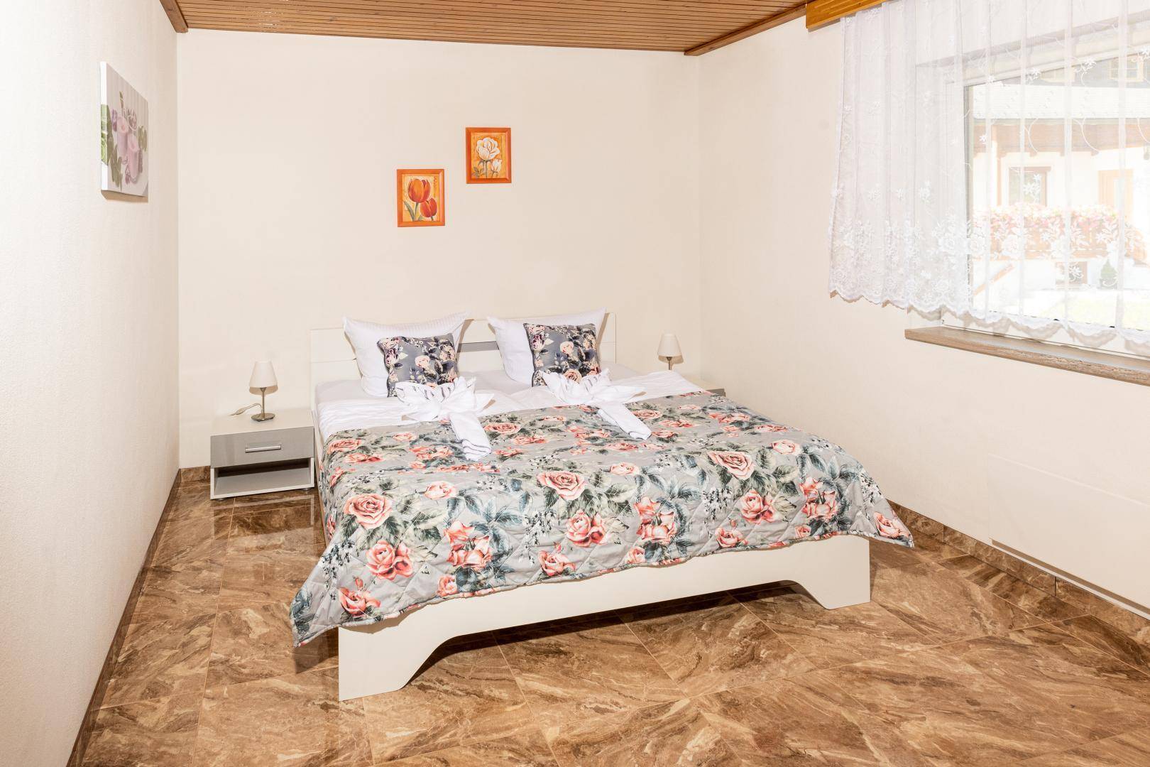Helles Schlafzimmer mit floraler Bettwäsche, Fliesenboden und Blick ins Grüne.