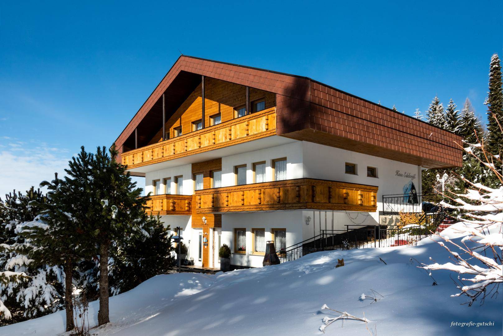 Alpines Haus im Winter, umgeben von Schnee und Bäumen, unter klarem blauem Himmel.