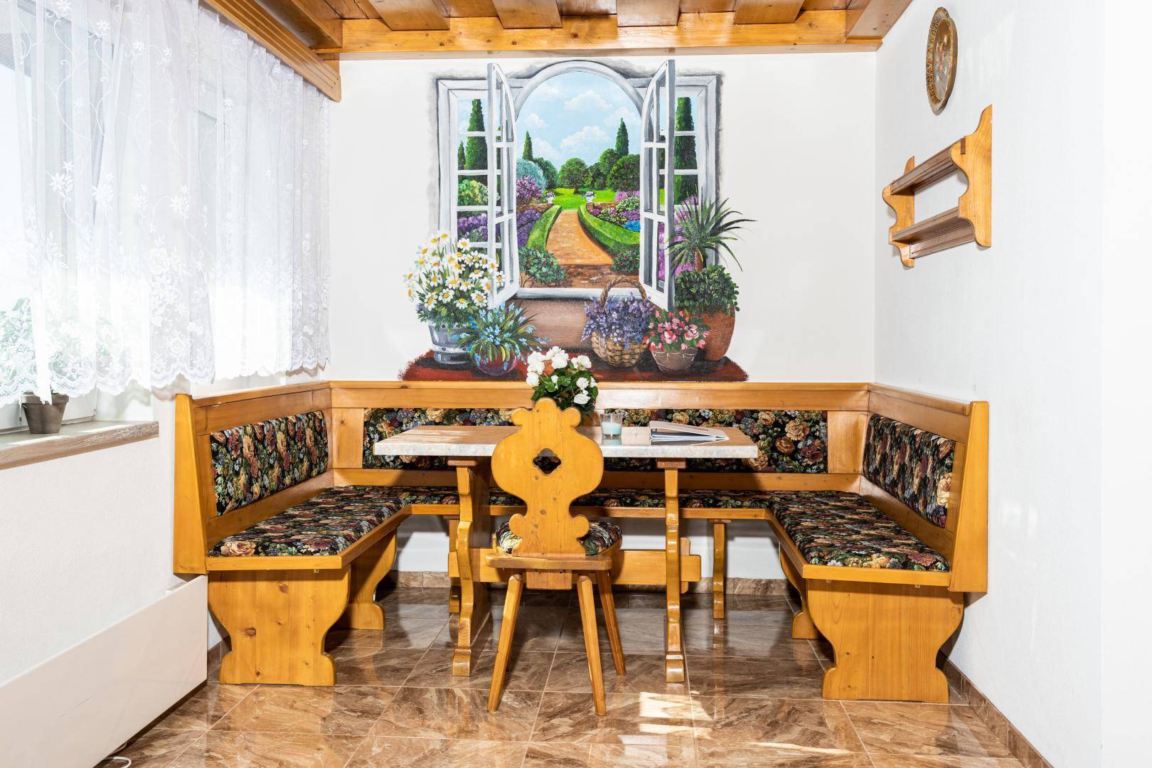Traditionelle Essecke im alpinen Stil mit Holzbänken, Fliesenboden und gemaltem Fensterbild.